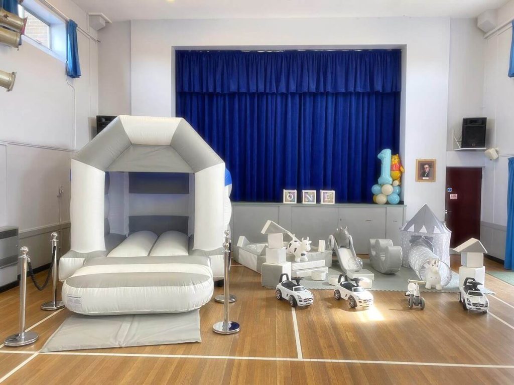 Bouncy castle and soft play equipment setup in a large hall with ride on cars and play tent.